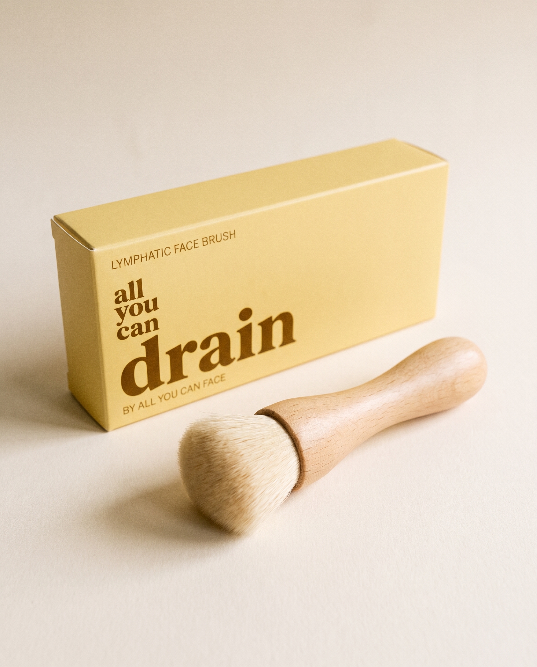 Eine weiche Bürste mit hellem Buchenholzgriff vor ihrer buttergelben Schachtel „all you can drain", auf sandfarbenem Papier.