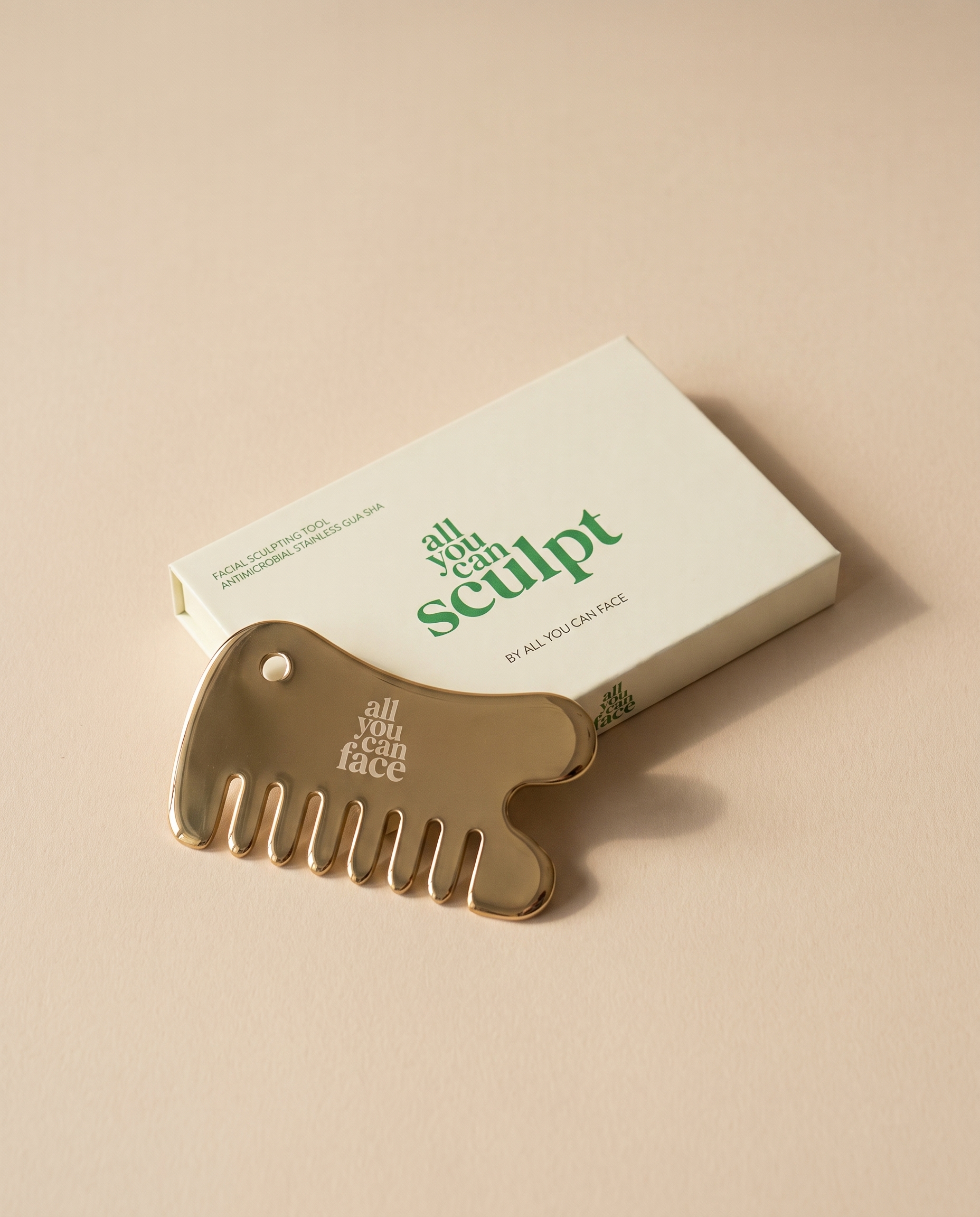 Der Kamm „all you can sculpt" aus poliertem, goldfarbenem Stahl, die Wortmarke in die Fläche graviert, an seiner cremefarbenen Schachtel auf sandfarbenem Papier.